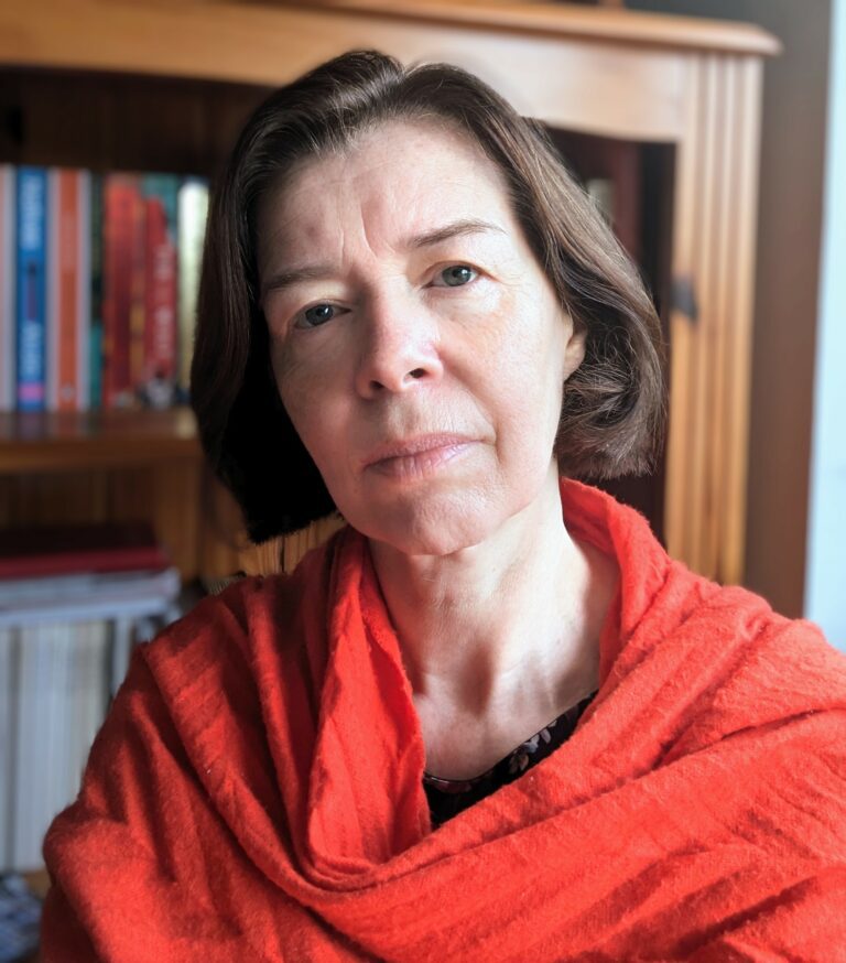 A white woman with a short, brunette bob and blue eyes is looking at the camera. She has a red scarf wrapped around her shoulders.