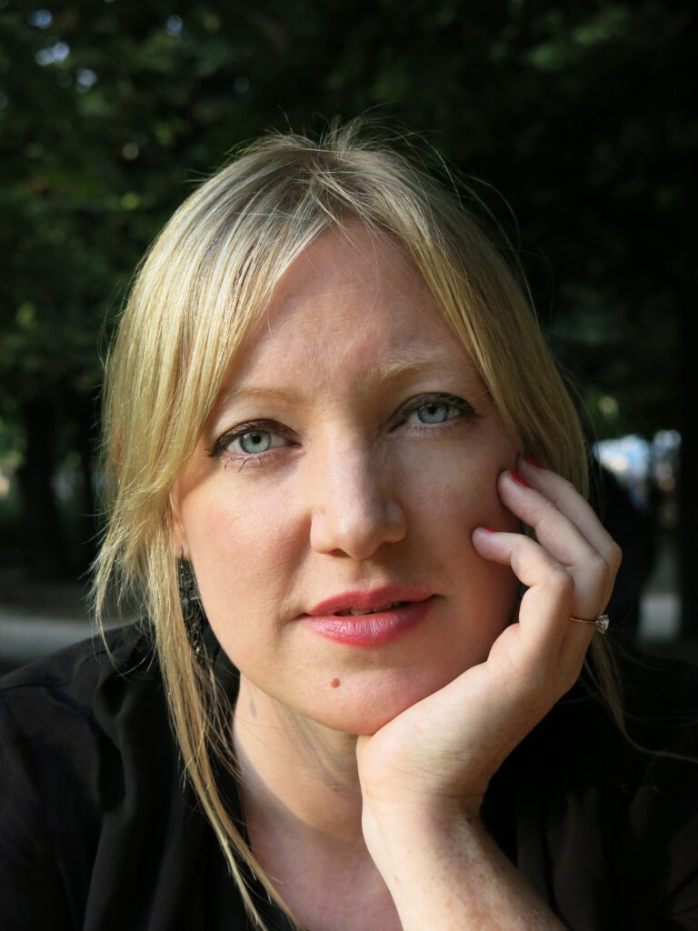 A blonde white woman with blue eyes is looking at the camera. Her hand cradles her face on one side.