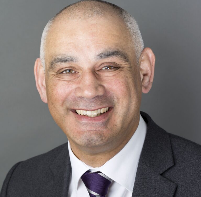 A bald male with brown eyes smiles at the camera. He looks friendly, and is wearing a suit.