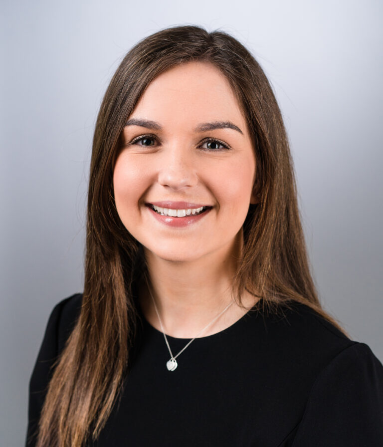 A brunette white lady is smiling at the camera. She has dark eyes and is wearing a heart shaped necklace.